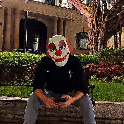 A front angled view of a person in a scary clown mask sitting on a bench in an outdoor setting